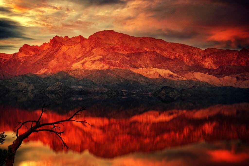 Vivid red‑orange sunset illuminating rugged mountains and their perfect reflection in a glassy lake.