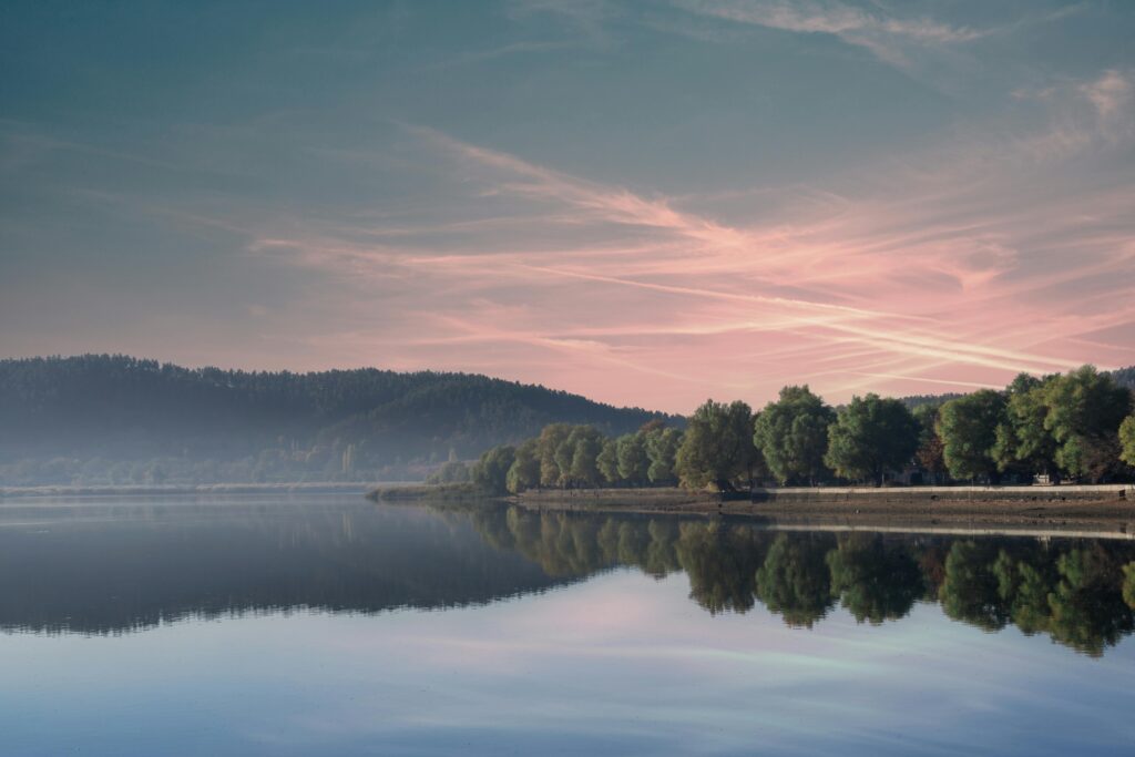 Serene landscape featuring a glassy lake, dense green treeline, and a softly coloured dawn sky.